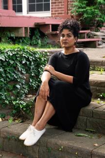 photo of divya victor, brown skin, black dress, white shoes, sitting on steps outside next a green bush.